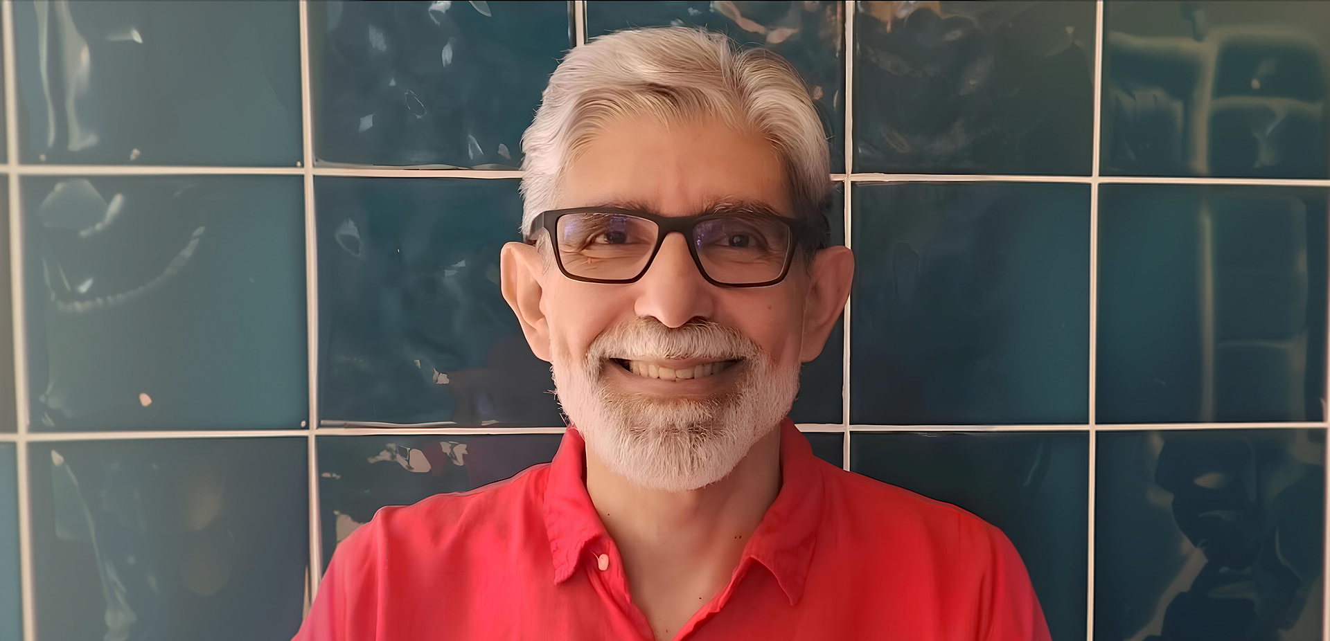 Um homem de cabelos e barba grisalhos, óculos pretos e camisa vermelha sorri para a câmera em frente a uma parede de azulejos verde-azulados.