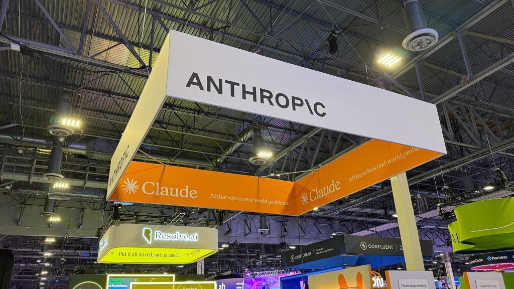 Um letreiro de estande suspenso em uma feira, exibindo "ANTHROPIC" em branco e "Claude AI that solves real-world problems" em laranja.