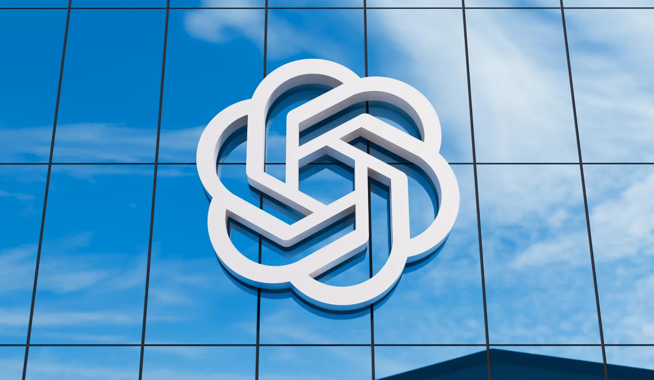 O logotipo branco e estilizado da OpenAI está fixado na fachada de vidro azul de um prédio moderno, com reflexos de nuvens.
