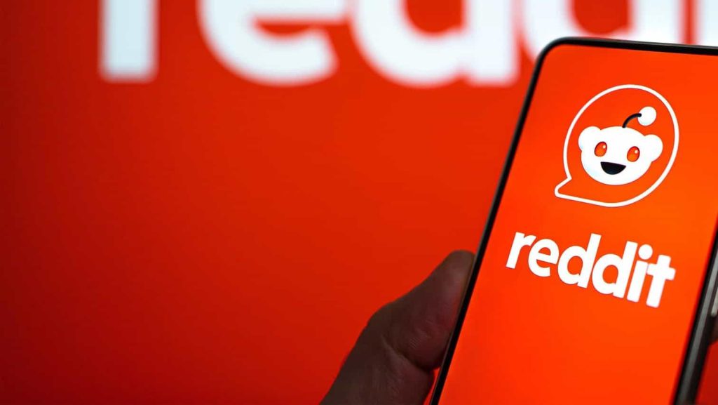 Uma mão segura um smartphone exibindo o aplicativo Reddit com o logotipo do Snoo e o nome reddit em um fundo vermelho desfocado.