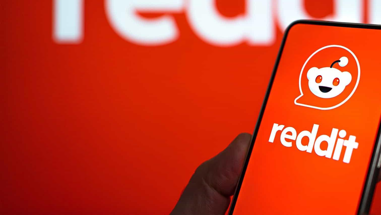 Uma mão segura um smartphone exibindo o aplicativo Reddit com o logotipo do Snoo e o nome reddit em um fundo vermelho desfocado.