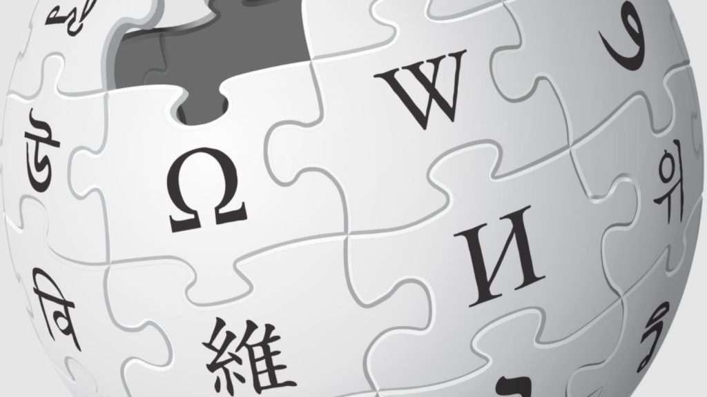 Um globo de quebra-cabeça branco e cinza, logotipo da Wikipédia, exibe caracteres de múltiplos idiomas com uma peça faltando no topo.