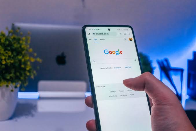 Uma mão segura um smartphone exibindo a página inicial do Google, com "Indonesia" e um dedo na tela, e um laptop com uma planta ao fundo.